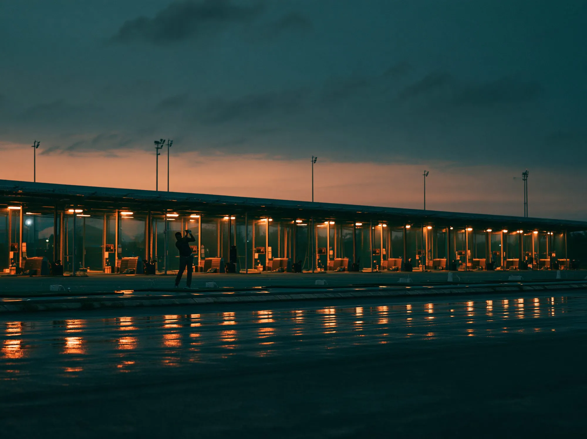 A driving range at dusk, bays lit warm against a darkening sky